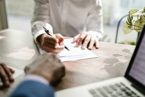 Person's hand writing on paper with a plant and laptop on the table - Financial Planner Image