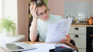 A woman looking at paper with glasses and a pen in hand; A laptop on the table with calculator and papers - How much interest you are paying - Wealth Connexion blog image