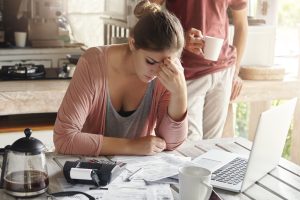 Thoughtful stressed young female sitting at kitchen table with papers and laptop computer trying to work through pile of bills, frustrated by amount of domestic expenses while doing family budget